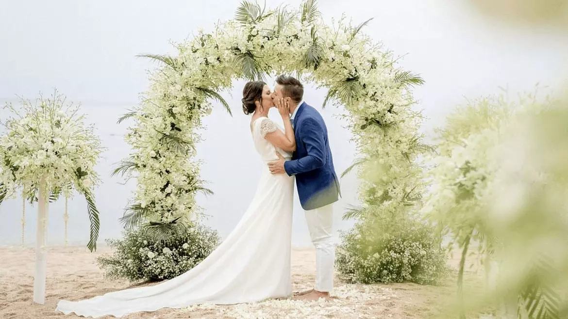 A couple kisses under a floral arch on the beach at Garrya Tongsai Bay Samui wedding venue.