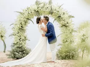 A couple kisses under a floral arch on the beach at Garrya Tongsai Bay Samui wedding venue.